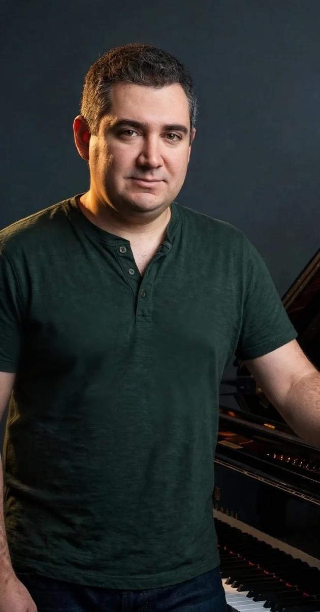 Jason Solowsky in a dark green henley shirt beside his black grand piano — his real photograph