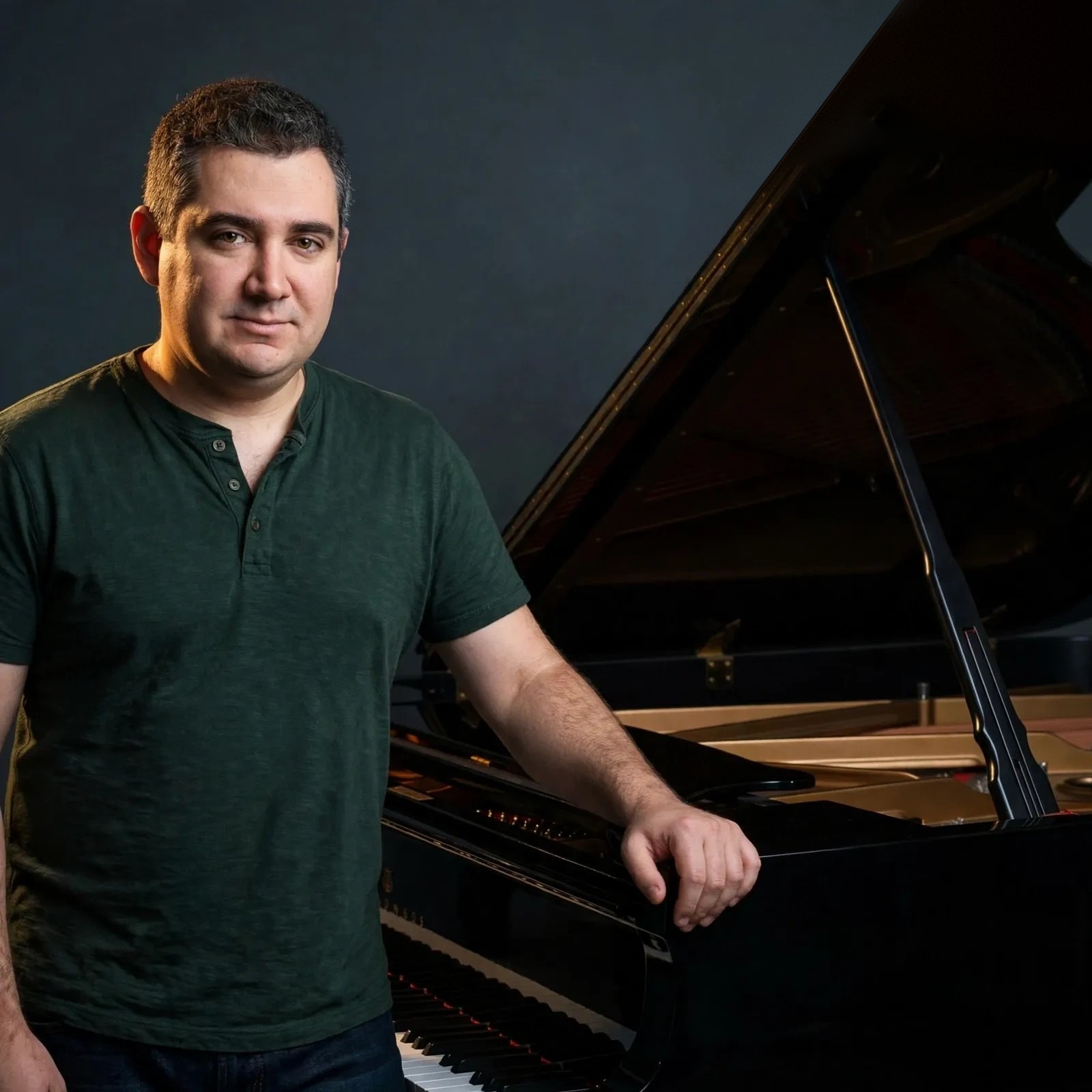 Jason Solowsky in a dark green henley standing beside his black grand piano with the lid raised, his hand resting on the piano — his real photograph, shown at its widest framing so the whole piano is visible